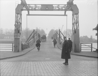 9 FDSPAARNE009 De brug te Deventer ligt hoog; fietsers en een paardenkar rijden tegen een steile brughelling op. Aan de ...
