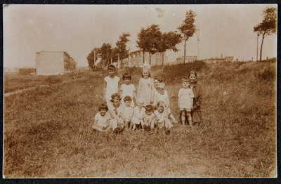102 Vrouw en 12 kinderen poseren in gras (aan een dijk?). Op de achtergrond laagbouwflats. Lokatie onbekend. Procedé: ...