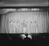 1324 Toneelvoorstelling: 7 dames in een rijtje op het toneel. Het doek is dicht, de pianist is zichtbaar in de orkestbak.