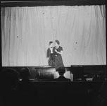 1341 Toneelvoorstelling: vrouw met buikspreekpop op het toneel, voor het dichte doek. Pianist is zichtbaar in de orkestbak.