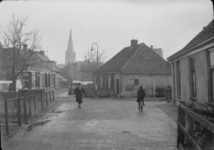 116 Wijhe: Opname van een straat in het dorp, met huizen, een voetganger en een fietser en een kerktoren op de ...